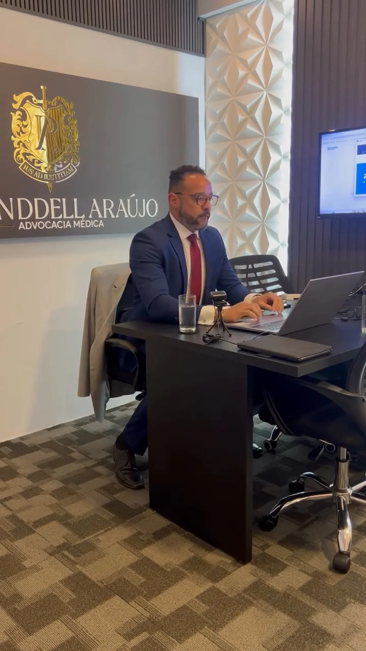 A sala do escritório: Wenddell Araújo, de terno azul, à mesa com o notebook e a câmera, sob a placa com o brasão dourado